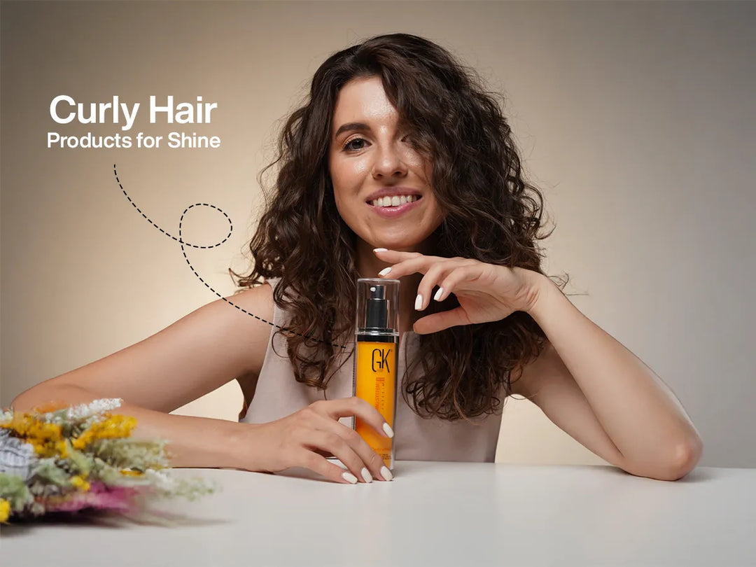 Woman with curly hair smiles while holding a bottle of GK Hair CurlDefineher on a table. Text reads "Curly Hair Products for Shine." Flowers in foreground.