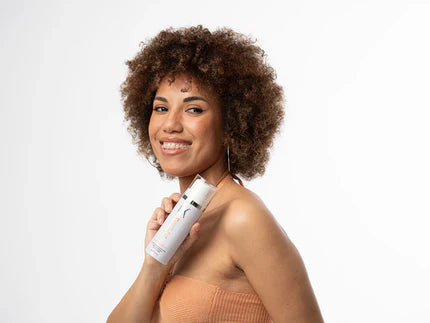 A person with curly hair holds a bottle of leave-in conditioner near their shoulder, wearing a strapless top against a white background.