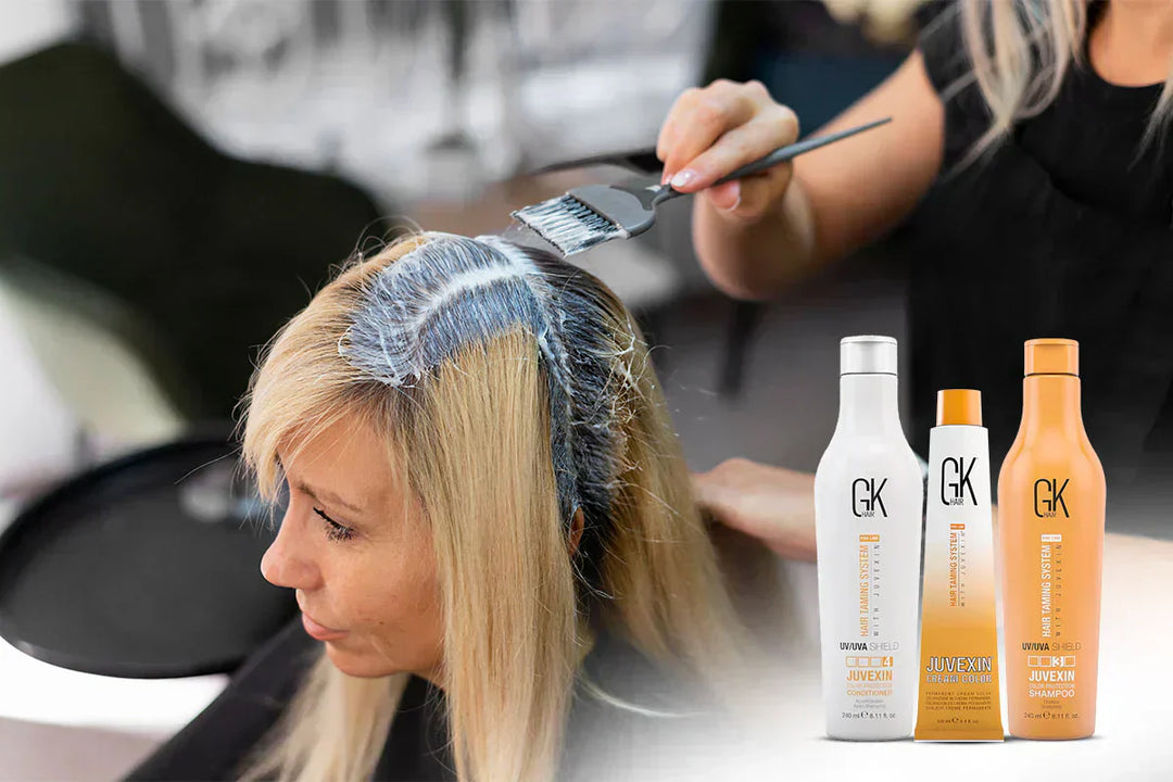 A hairstylist applies color to blonde hair using a brush, with hair care products displayed nearby.
