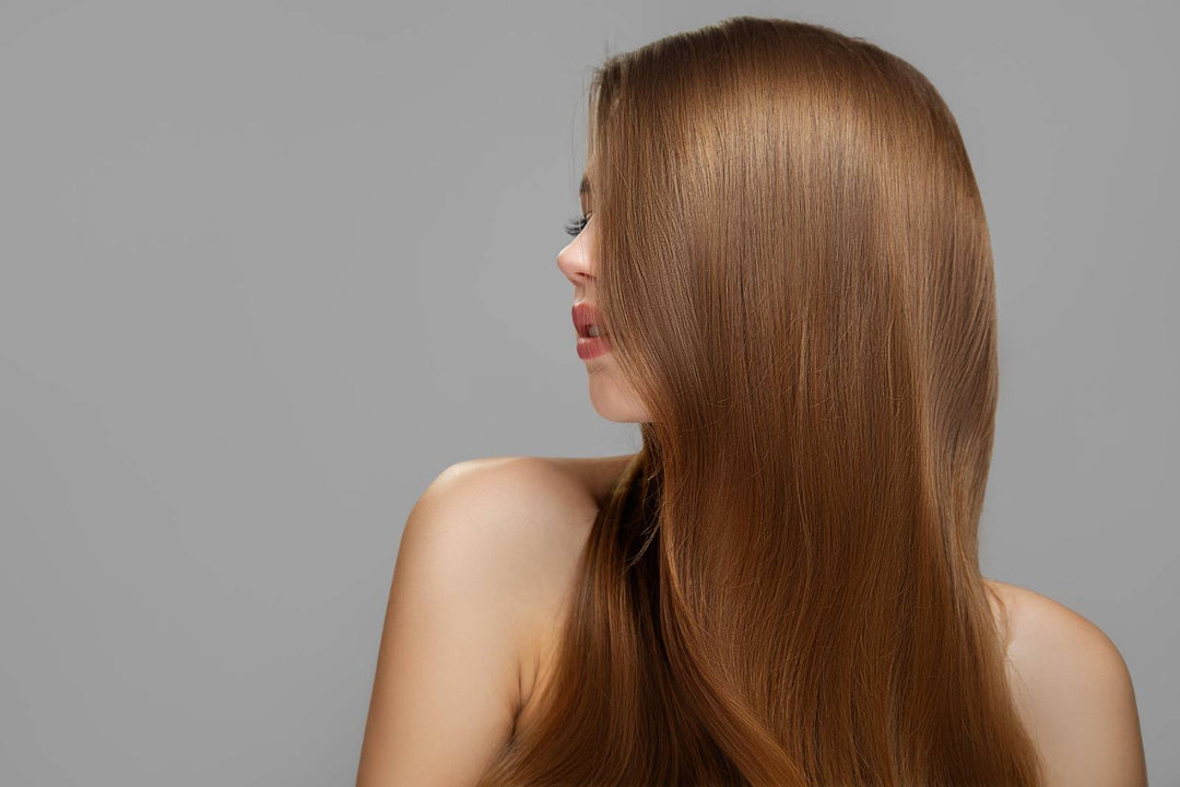 A woman with long, shiny, Brown hair turned sideways, showcasing smooth and healthy strands against a neutral gray background.